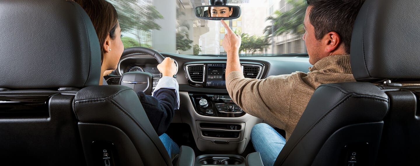 An interior view of a 2021 Chrysler Pacifica Limited with the driver focusing on the road ahead and the front passenger pressing a button on the rearview mirror.