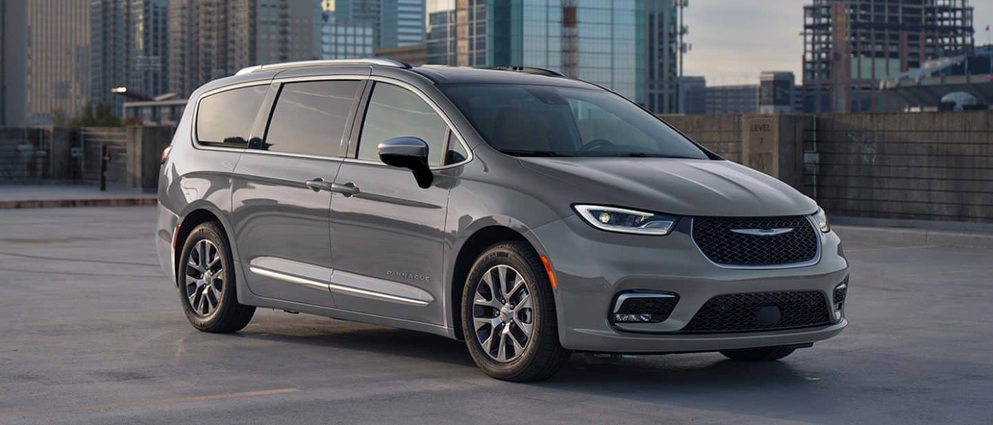 Display A passenger-side front angle of a gray 2025 Chrysler Pacifica Pinnacle Plug-in Hybrid parked on the roof of a parking structure with tall buildings in the background.