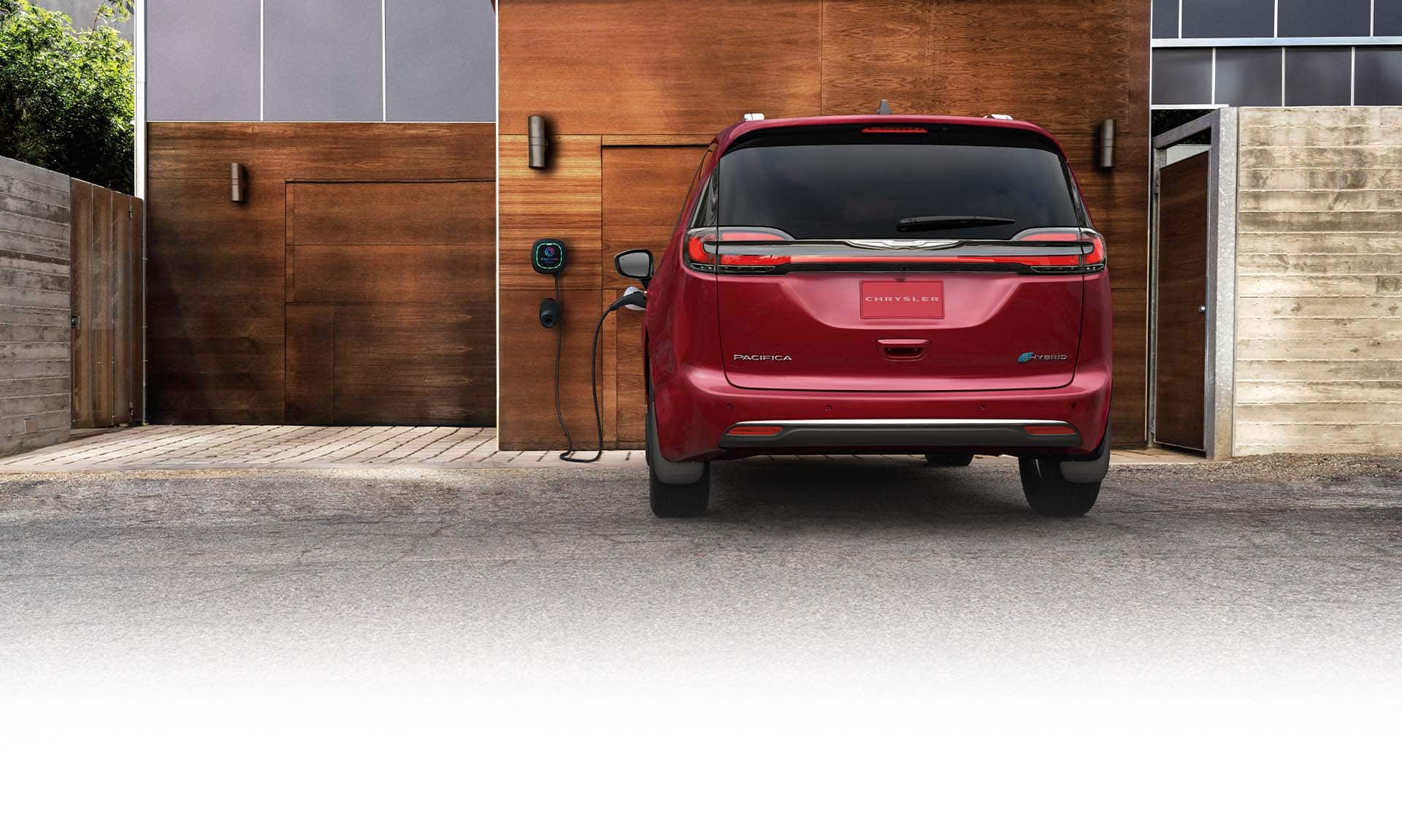 A rear angle of a red 2025 Chrysler Pacifica Pinnacle Plug-In Hybrid parked outside a garage that has a built-in Level 2 charging station, with a charging cord plugged into the vehicle's charging port.