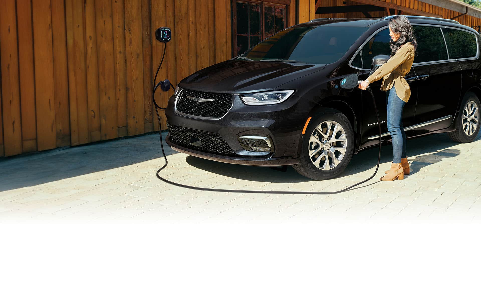 A driver-side front angle of a black 2025 Chrysler Pacifica Pinnacle Plug-in Hybrid parked outside a building with a charging station. A woman is plugging the station's charging cord into the vehicle's charging port.